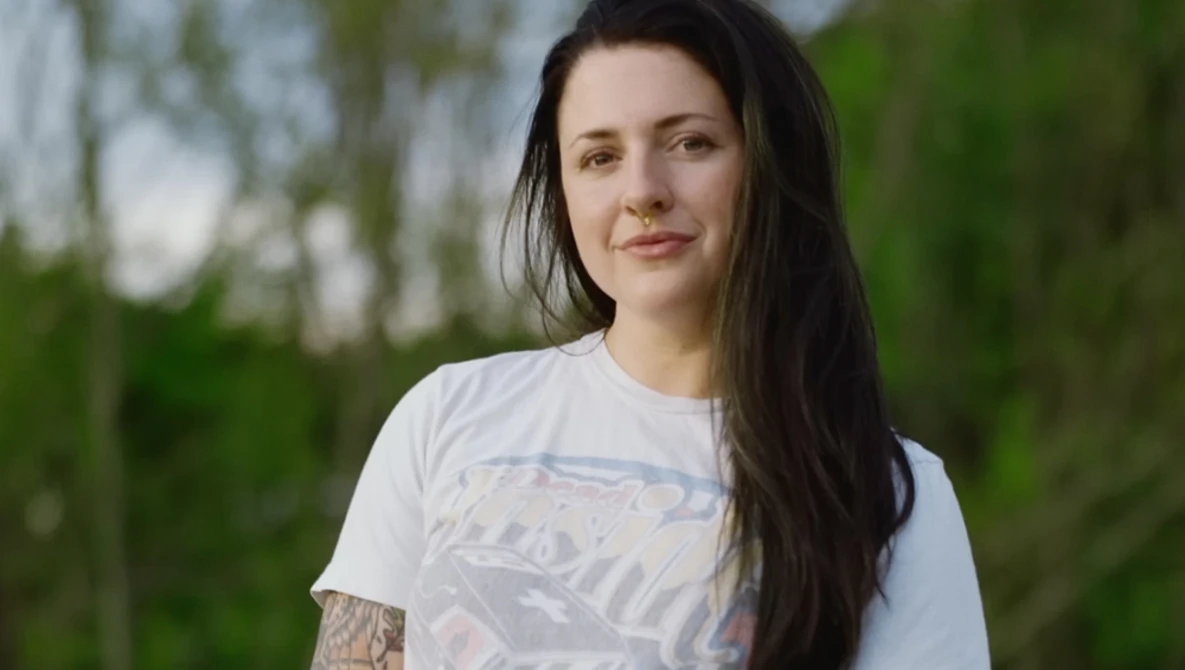 Woman with long dark hair wearing a white graphic t-shirt, photographed outdoors with soft natural light.