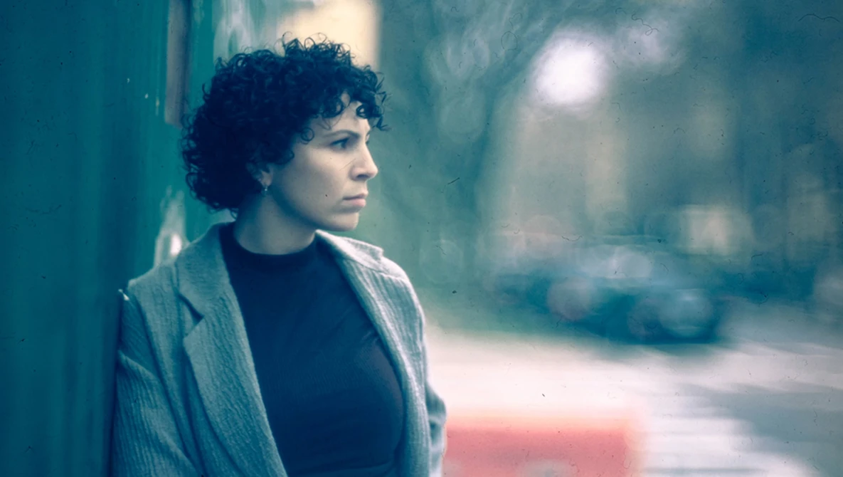Person with curly dark hair wearing a cardigan over a dark shirt, gazing sideways in an urban alley.