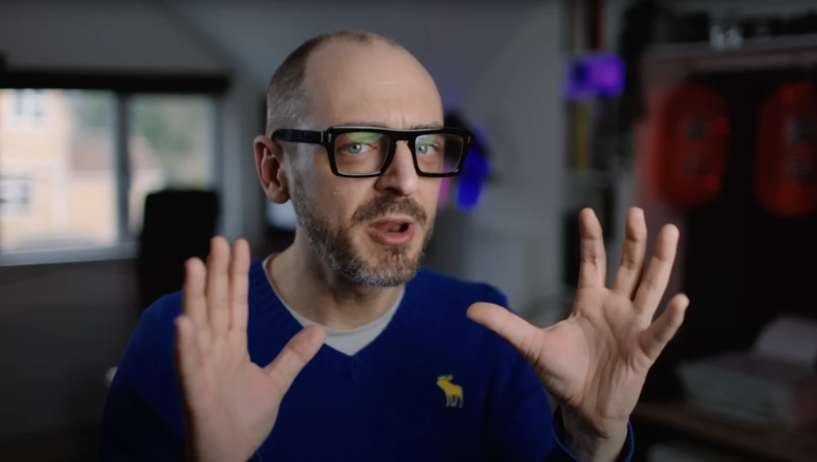 Man with glasses wearing navy blue shirt gestures expressively while speaking in studio setting.