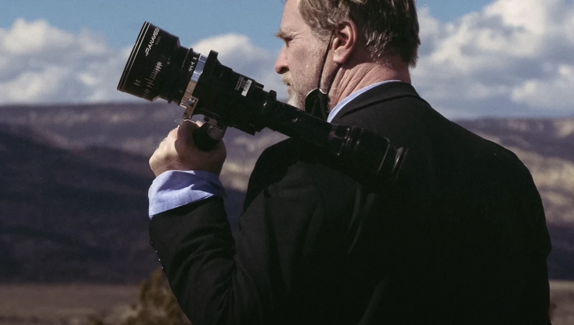 Photographer in dark jacket holding a telephoto lens to his eye, photographing a vast canyon landscape under blue sky.