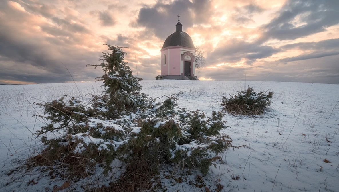 Snow-covered landscape with a domed chapel and bare shrubs under dramatic cloudy sky.