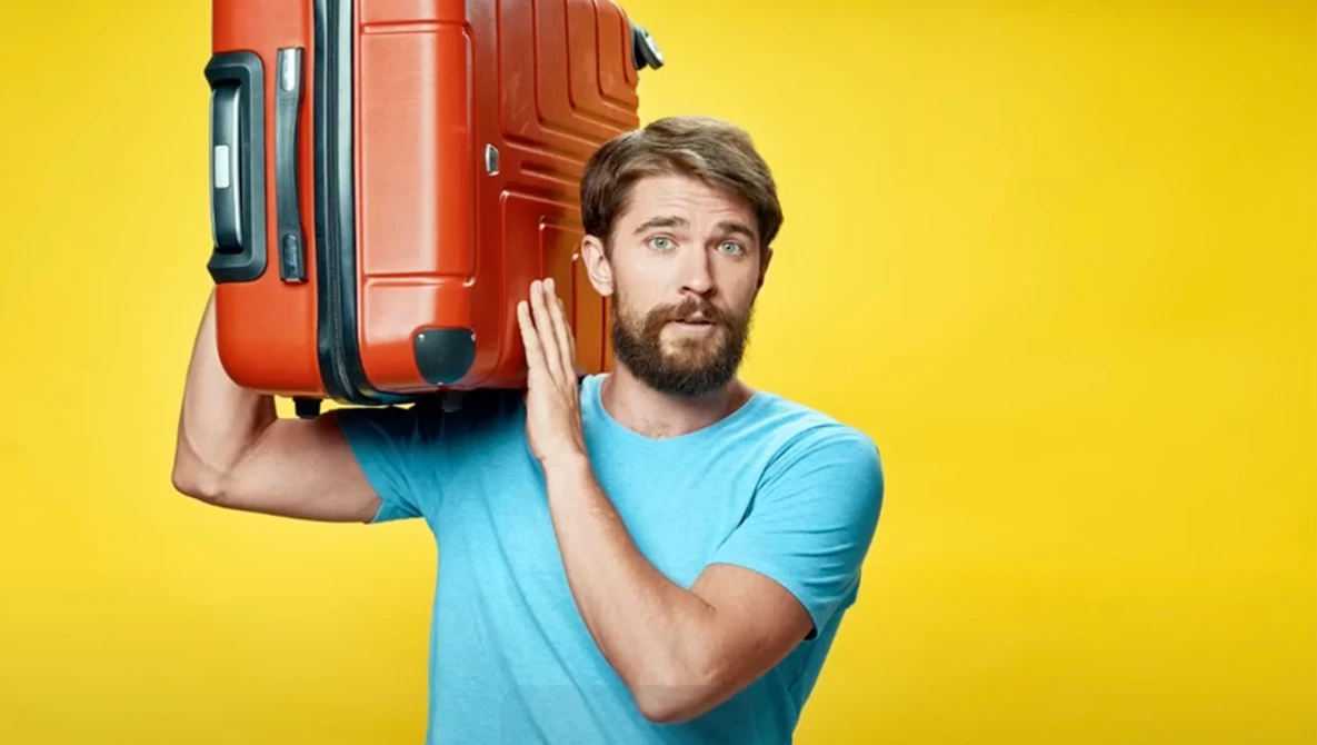 Bearded man in light blue t-shirt carrying red suitcase on shoulder against yellow background.