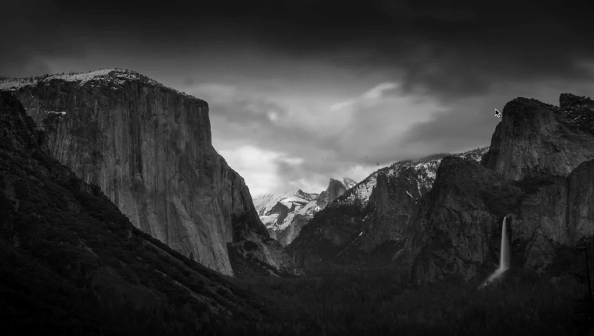 Black and white landscape of granite cliffs framing a valley with a waterfall and snow-capped peaks in the distance.