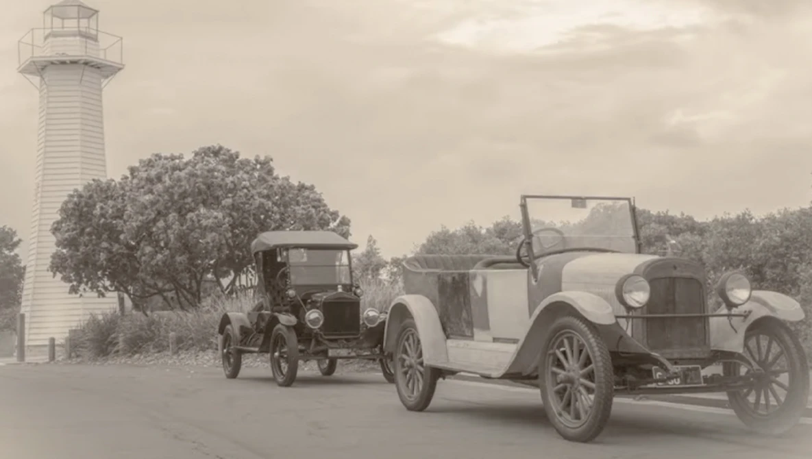 Two vintage automobiles parked near a lighthouse under overcast sky.