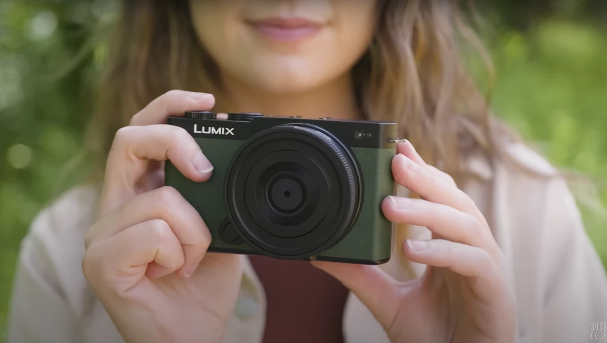 Woman holding a black Lumix compact camera outdoors with blurred green background.