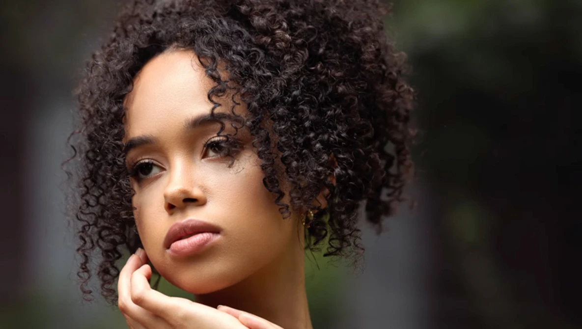 Portrait of a young woman with dark curly hair, looking toward the camera with a contemplative expression.