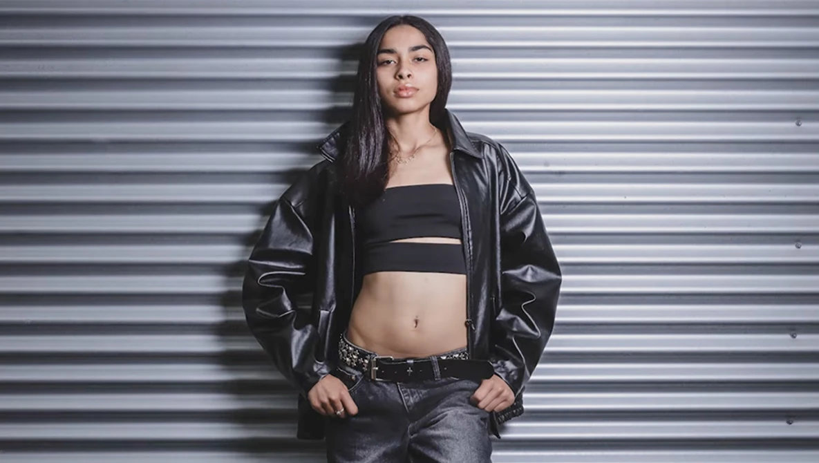 Woman in black leather jacket and crop top posing against corrugated metal wall.