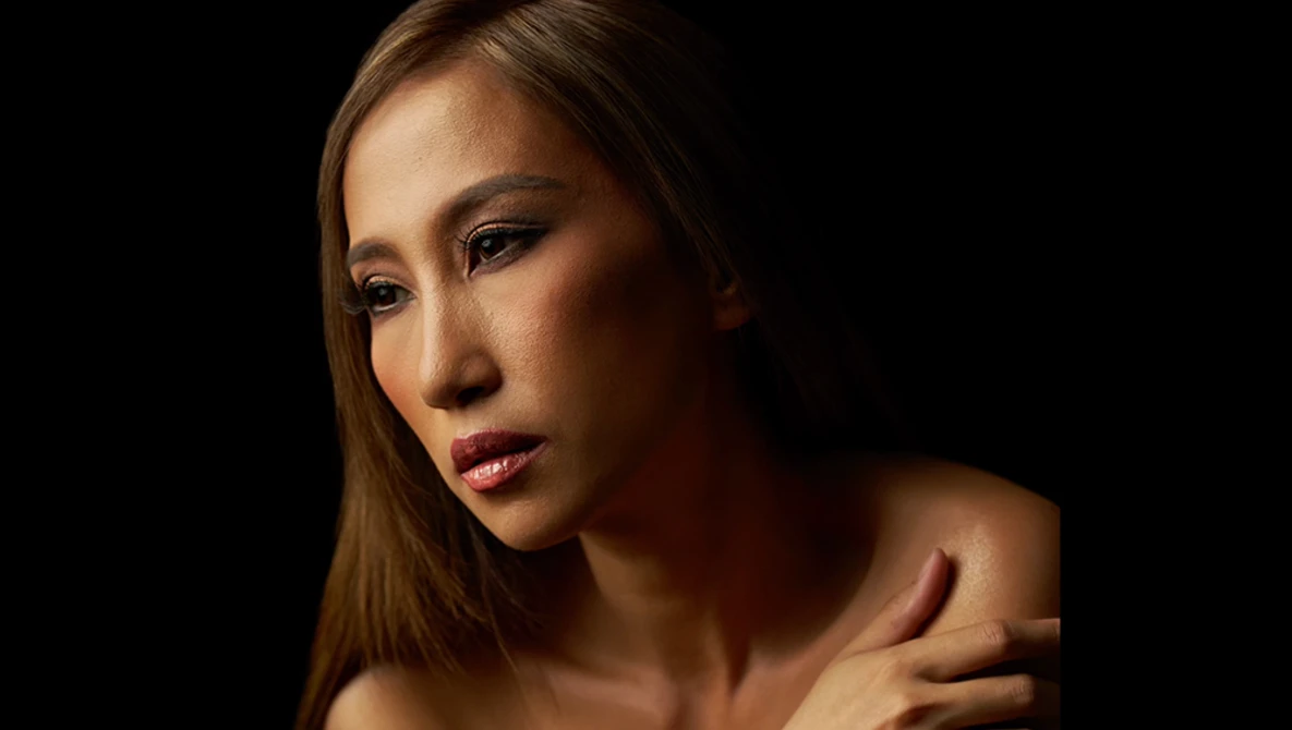 Woman with long brown hair gazes downward against a black background with dramatic side lighting.