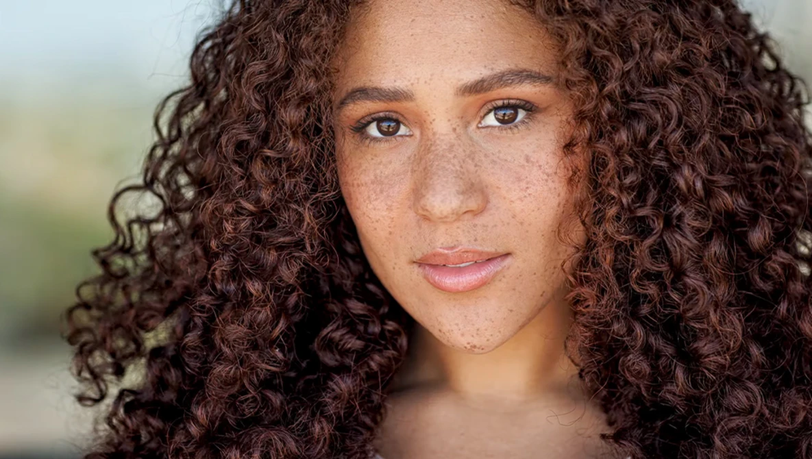 Portrait of a woman with long curly brown hair and freckles looking directly at camera.