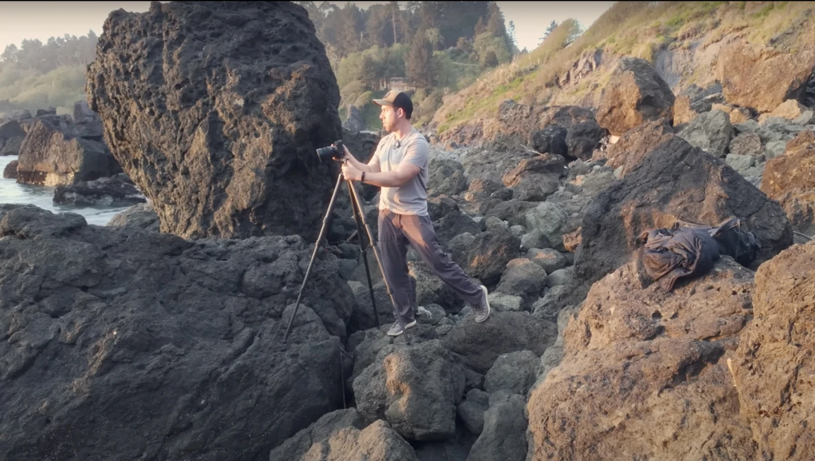 Photographer on tripod positioning among rocky coastal formations with water and forested hills in background.
