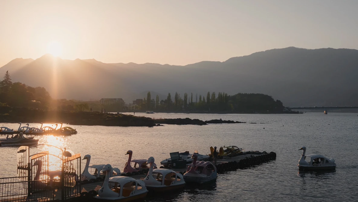 Golden sunrise light illuminates a calm alpine lake with moored boats in the foreground and mountains silhouetted across the water.