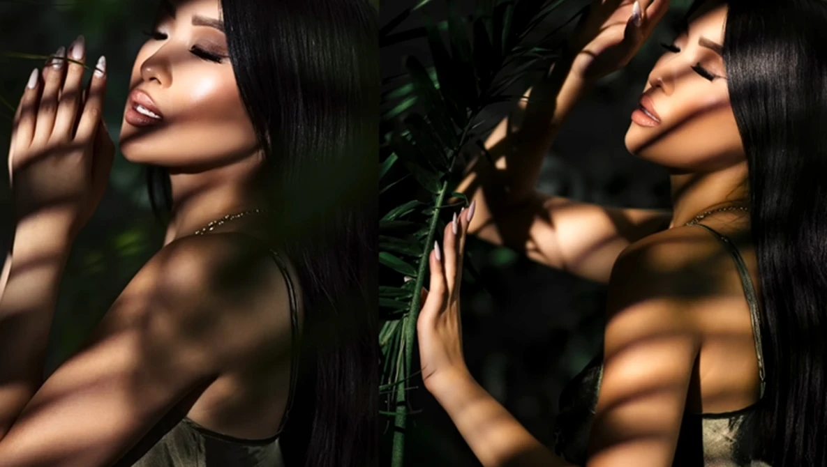 Woman with long black hair posing among tropical foliage with dappled sunlight and shadows across her face and shoulders.