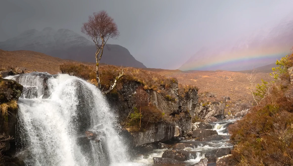 Waterfall cascades over rocky terrain with a solitary tree on the hillside and a rainbow arcing across the misty valley beyond.