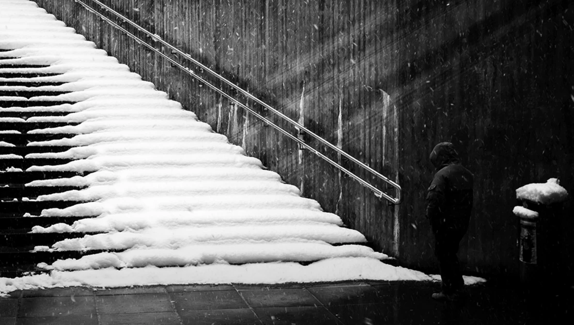 Person standing against a dark wooden wall beside a snow-covered wooden structure or bench.