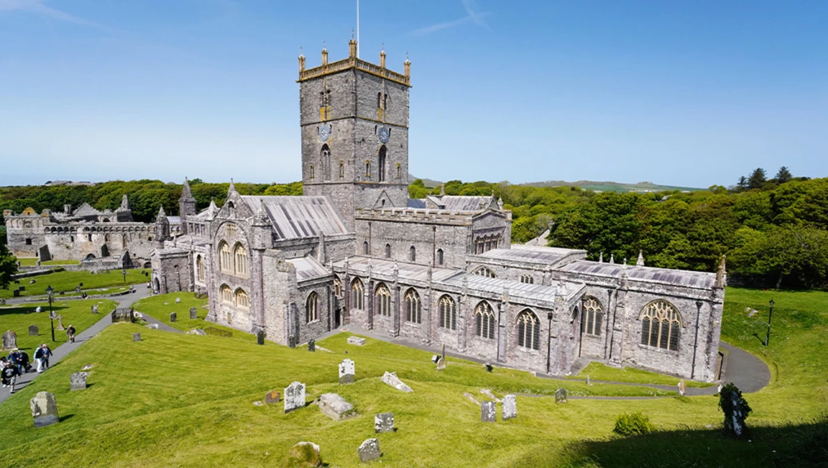 Medieval stone church with square tower and graveyard surrounded by green fields and trees under blue sky.