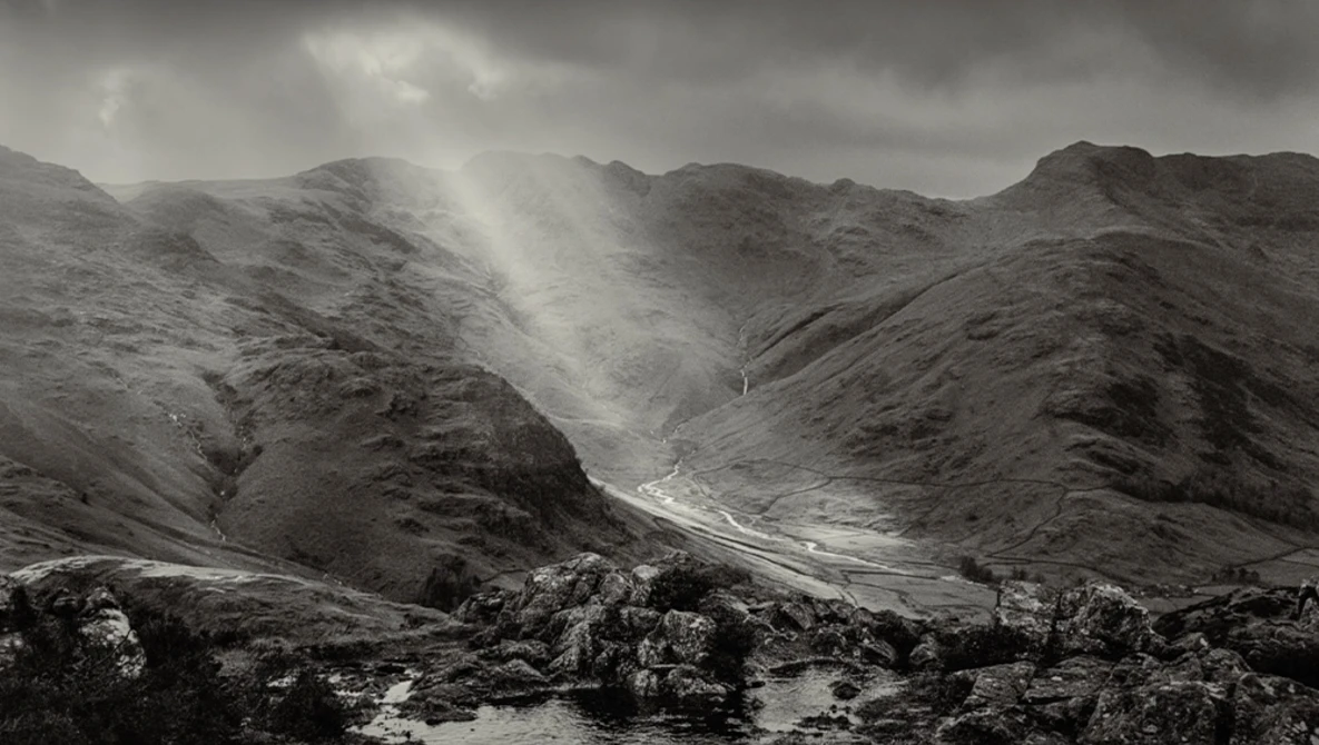 Dramatic mountain valley with crepuscular rays breaking through storm clouds over a winding river.