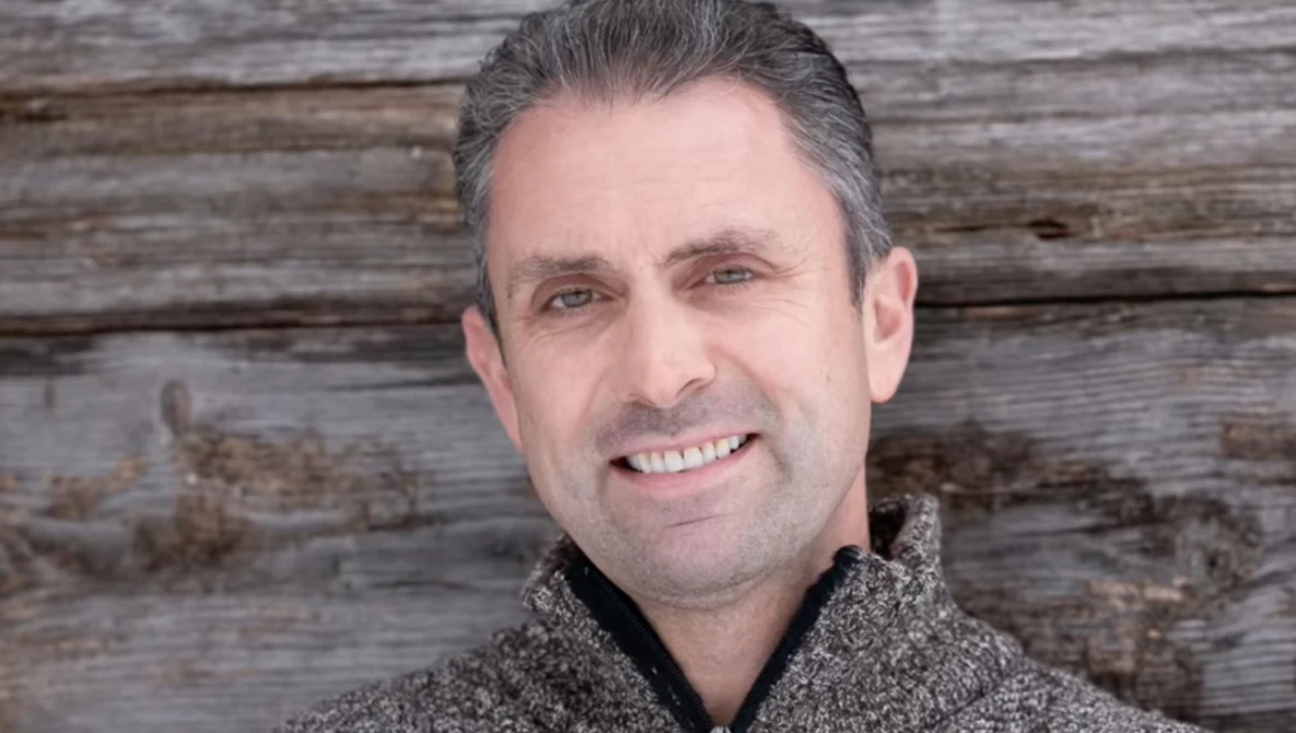 Man smiling at camera wearing gray knit sweater against weathered wood backdrop.