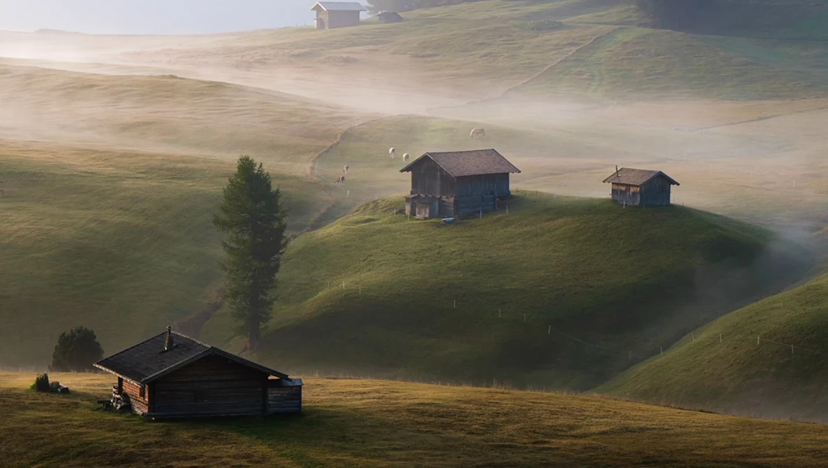 Misty alpine valley with scattered wooden chalets nestled among rolling green hills at golden hour.