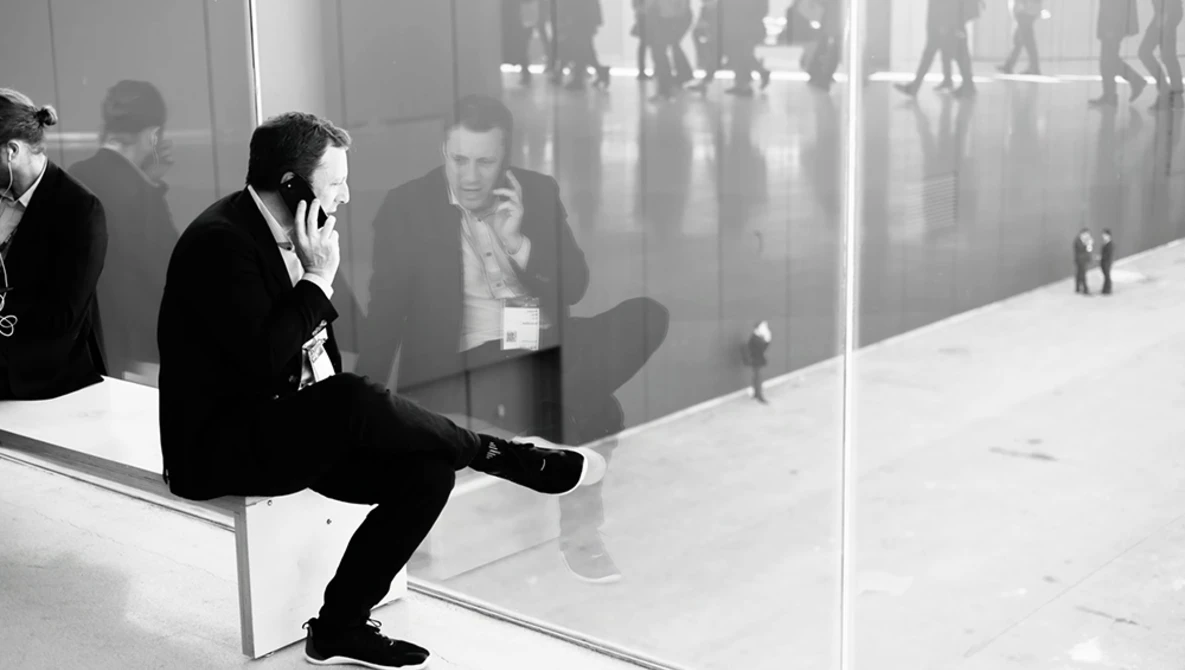 Businessman in dark suit sitting on white bench against glass window, speaking on phone while observing busy corridor reflection.