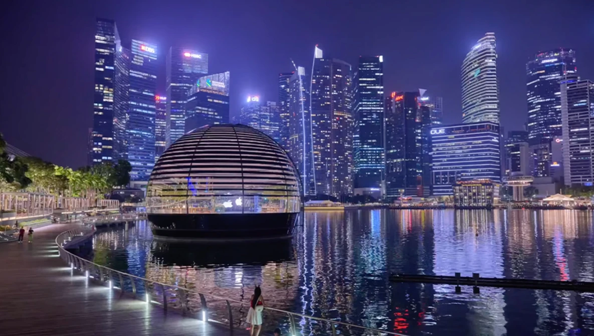 Modern dome structure on waterfront promenade with downtown city skyline reflected in calm water at night.