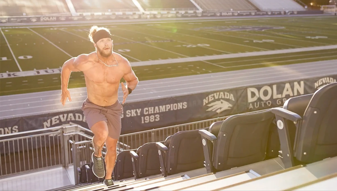 Shirtless runner in motion on a stadium press box with field and signage visible behind.