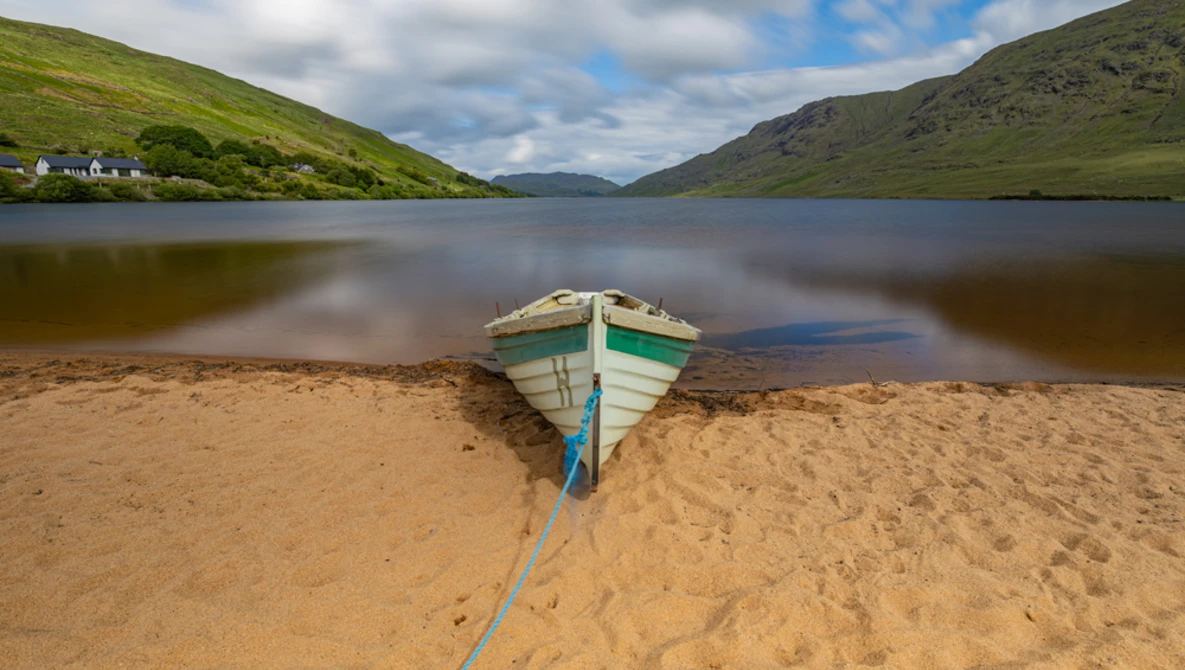 Blue and green rowboat beached on sand with a rope, facing calm water and green mountains under partly cloudy sky.