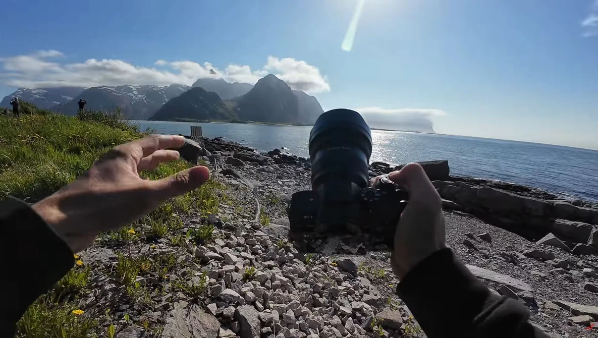 Photographer's hands holding a camera on a rocky beach with snow-capped mountains across the water.