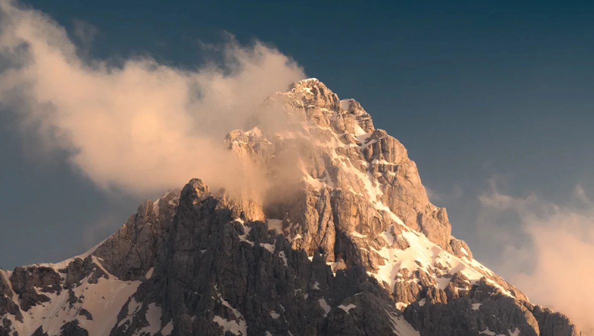 Snow-capped mountain peak bathed in golden light with clouds swirling around its summit.