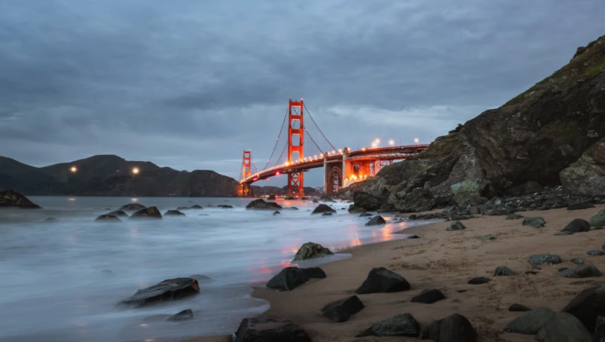 Golden Gate Bridge illuminated at dusk, viewed from a rocky beach with smooth water and dark boulders in foreground.