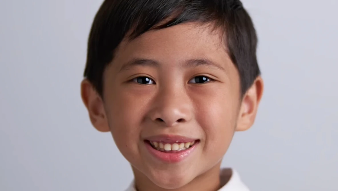 Young boy with dark hair smiling at camera against neutral gray background.