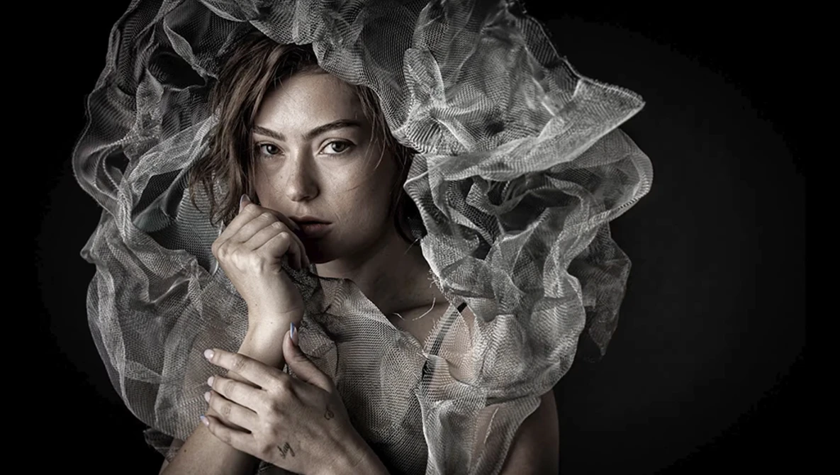 Woman with sheer draped fabric around her head and shoulders against a dark background.