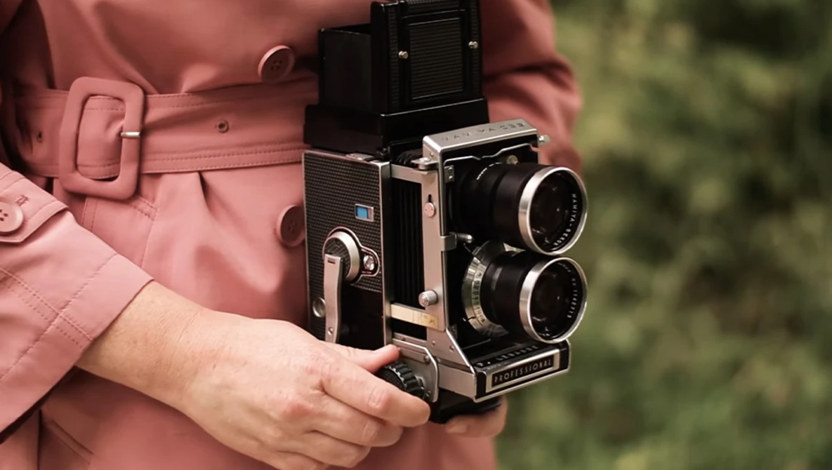 Person in rose-colored jacket holding a twin-lens reflex camera with dual objectives and viewfinder.