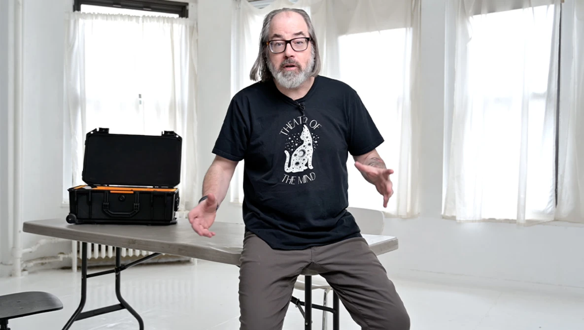 Man in glasses and black t-shirt sitting on table next to open equipment case in bright studio room.