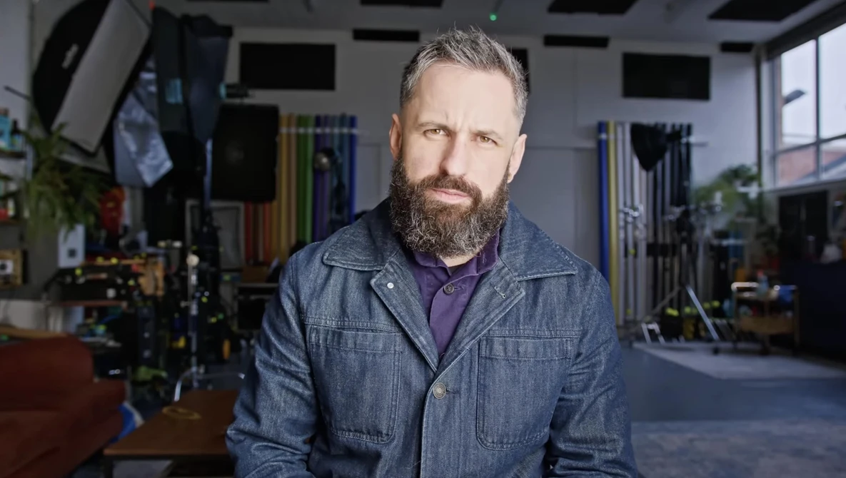 Man with full beard wearing denim jacket in professional studio setting with lighting equipment visible in background.