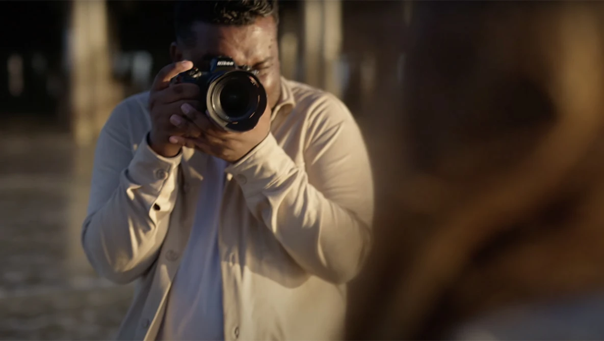 Photographer holding a DSLR camera with telephoto lens, shooting toward the camera in golden hour light.