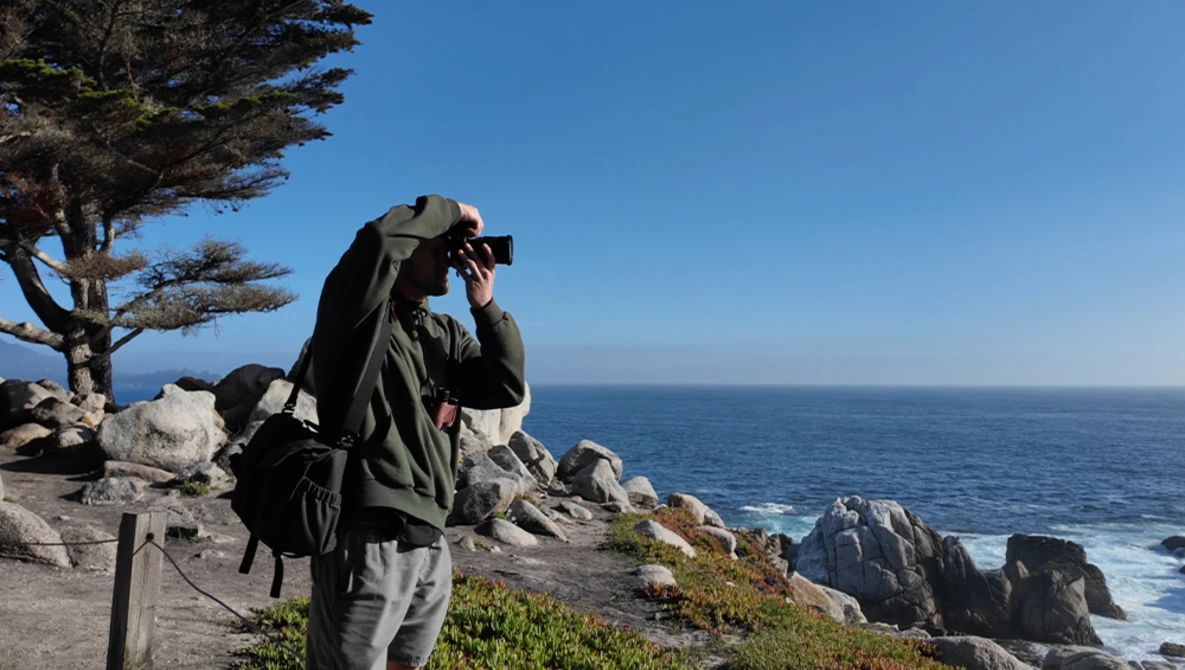 Photographer with camera and backpack on coastal rocks overlooking the ocean.