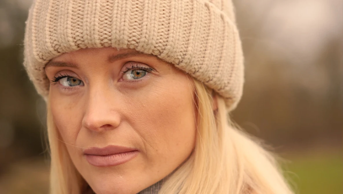 Woman with blonde hair wearing a cream knit beanie, photographed outdoors with soft natural lighting.