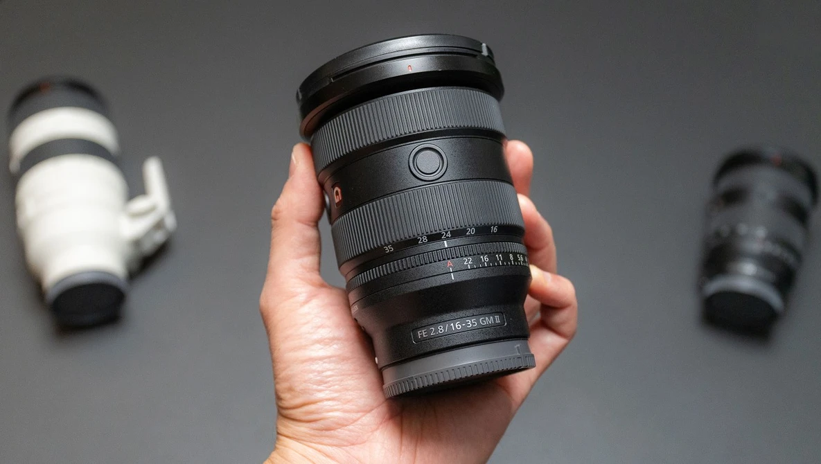 Hand holding a telephoto zoom lens with focus ring and depth-of-field scale visible against gray background.