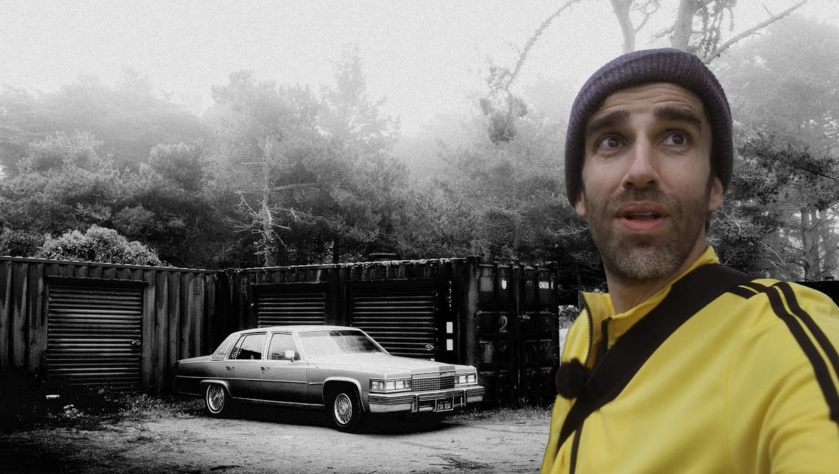 Man in yellow jacket and beanie looking upward beside a vintage car and industrial containers in misty forest setting.