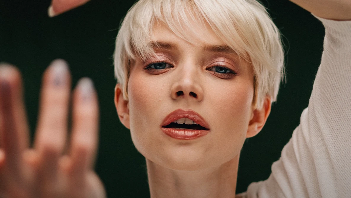 Portrait of a person with platinum blonde pixie cut against a dark background, with hands positioned near their face and shoulder.