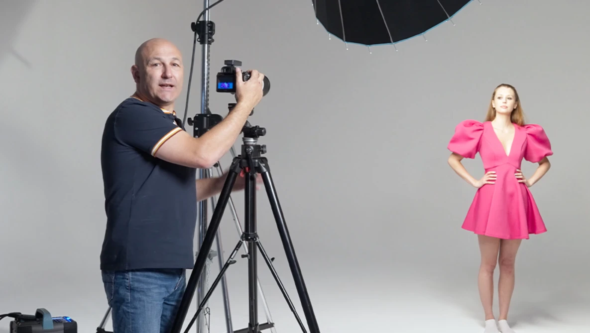 Photographer adjusting camera settings on tripod while model in pink dress poses under studio lighting.