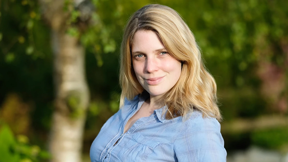 Woman with blonde shoulder-length hair wearing a light blue button-up shirt, photographed outdoors with soft natural lighting.