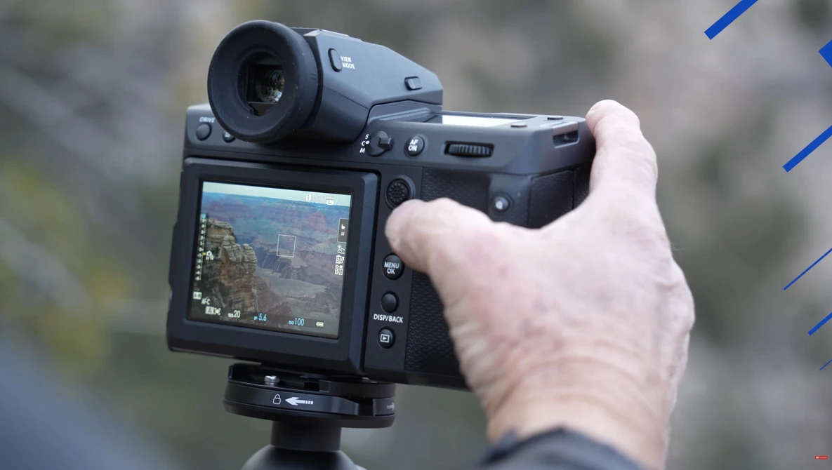 Hand holding a mirrorless camera with articulating LCD screen displaying live view of a landscape scene.