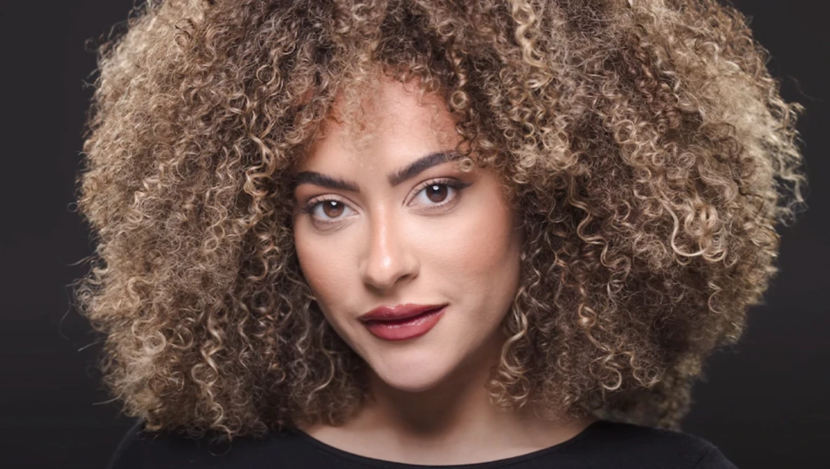 Portrait of a woman with voluminous curly blonde hair against a dark background.