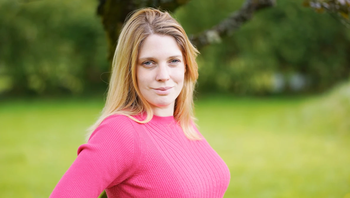 Woman with shoulder-length blonde hair wearing a pink ribbed sweater, photographed outdoors against a blurred green background.