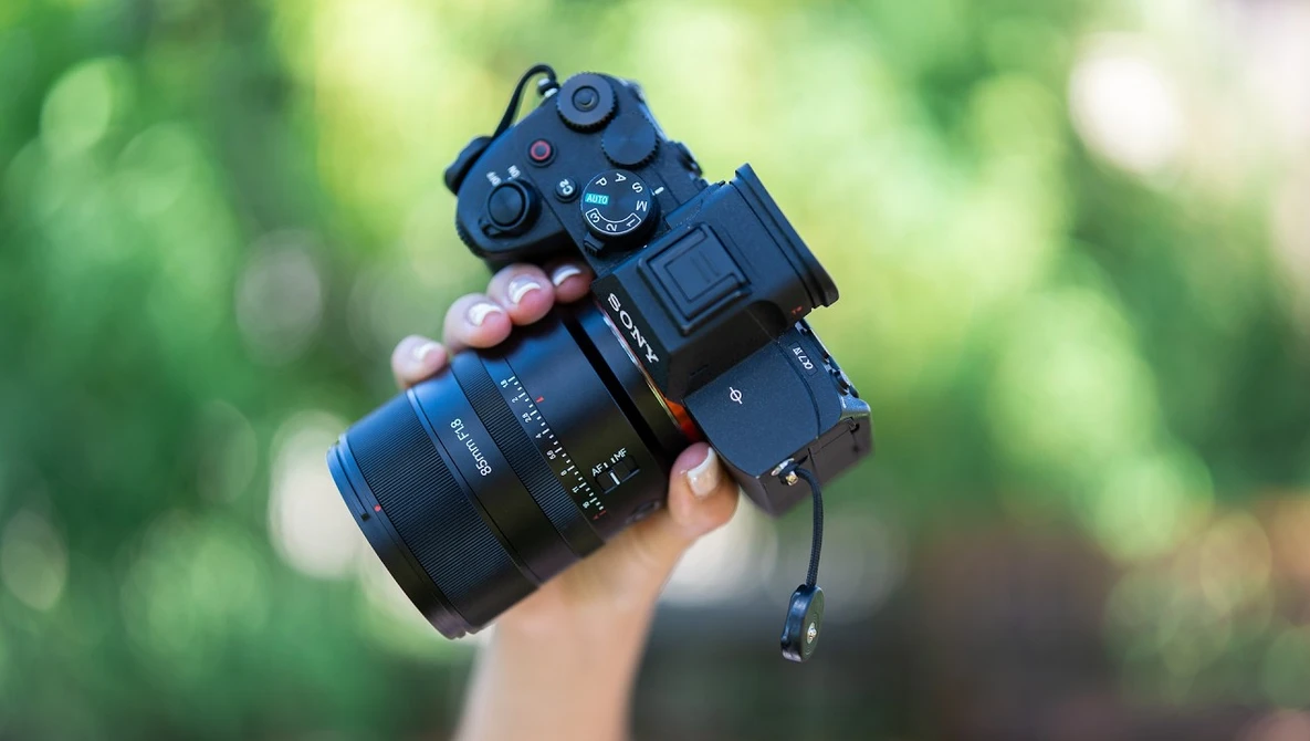 Photographer holding a mirrorless camera with attached lens and external recorder against a blurred green foliage background.