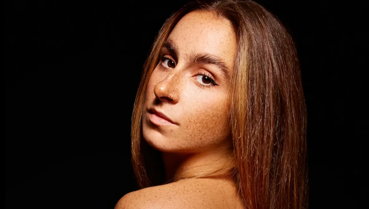 Woman with long brown hair looking over shoulder against black background.