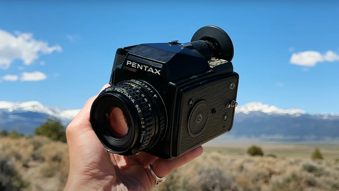 A Pentax 67 medium format camera held in someone's hand against a mountain landscape backdrop.
