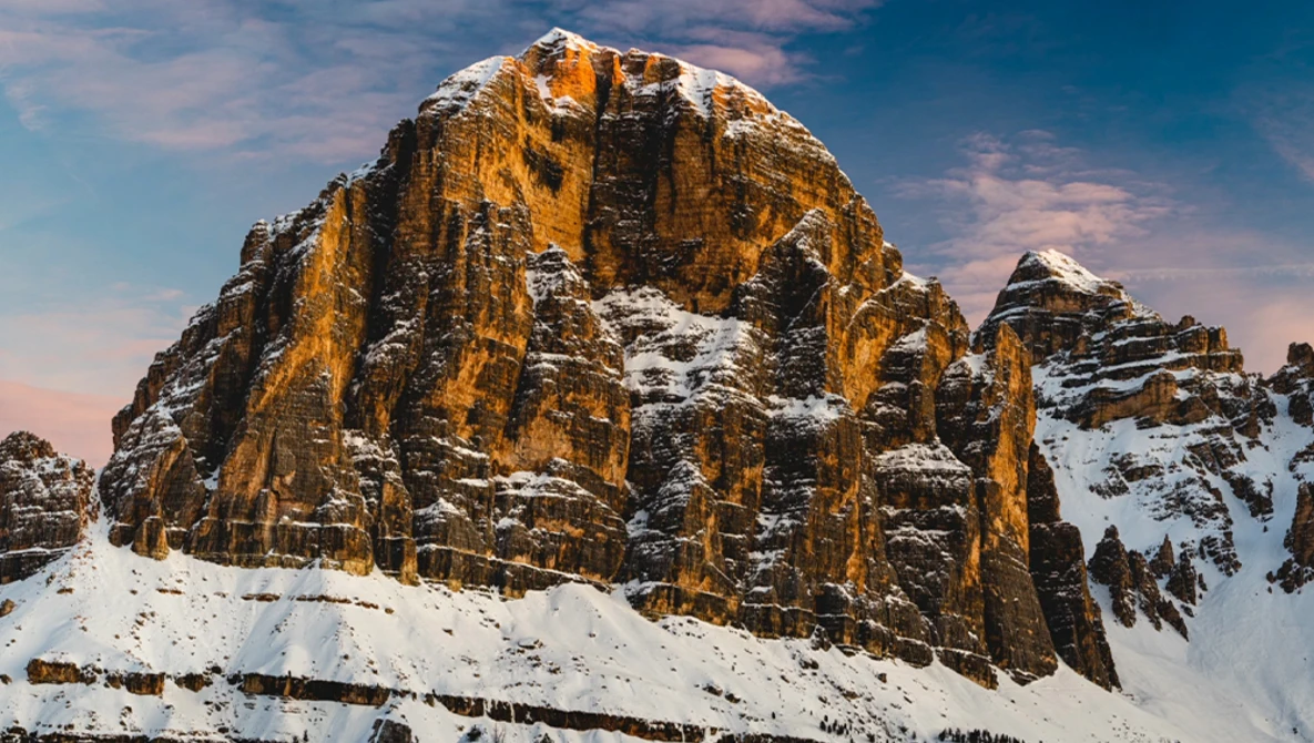 Snow-covered rocky mountain peak with golden-orange columnar formations lit by warm sunlight against blue sky.