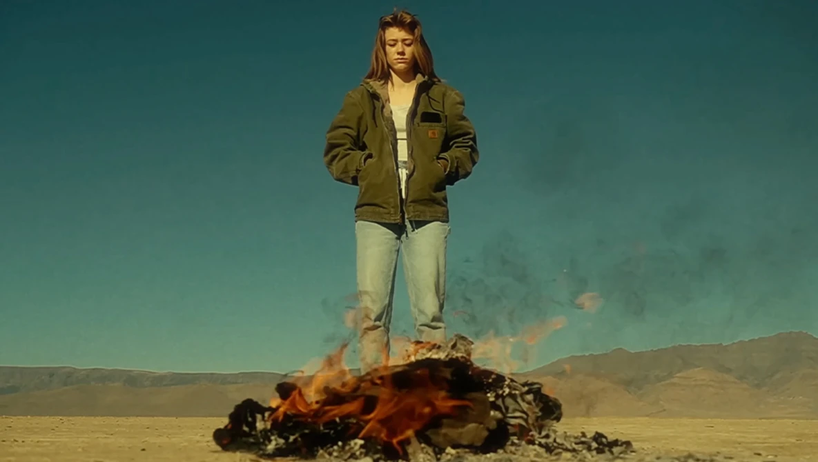 Person standing on burning debris in desert landscape under teal sky.